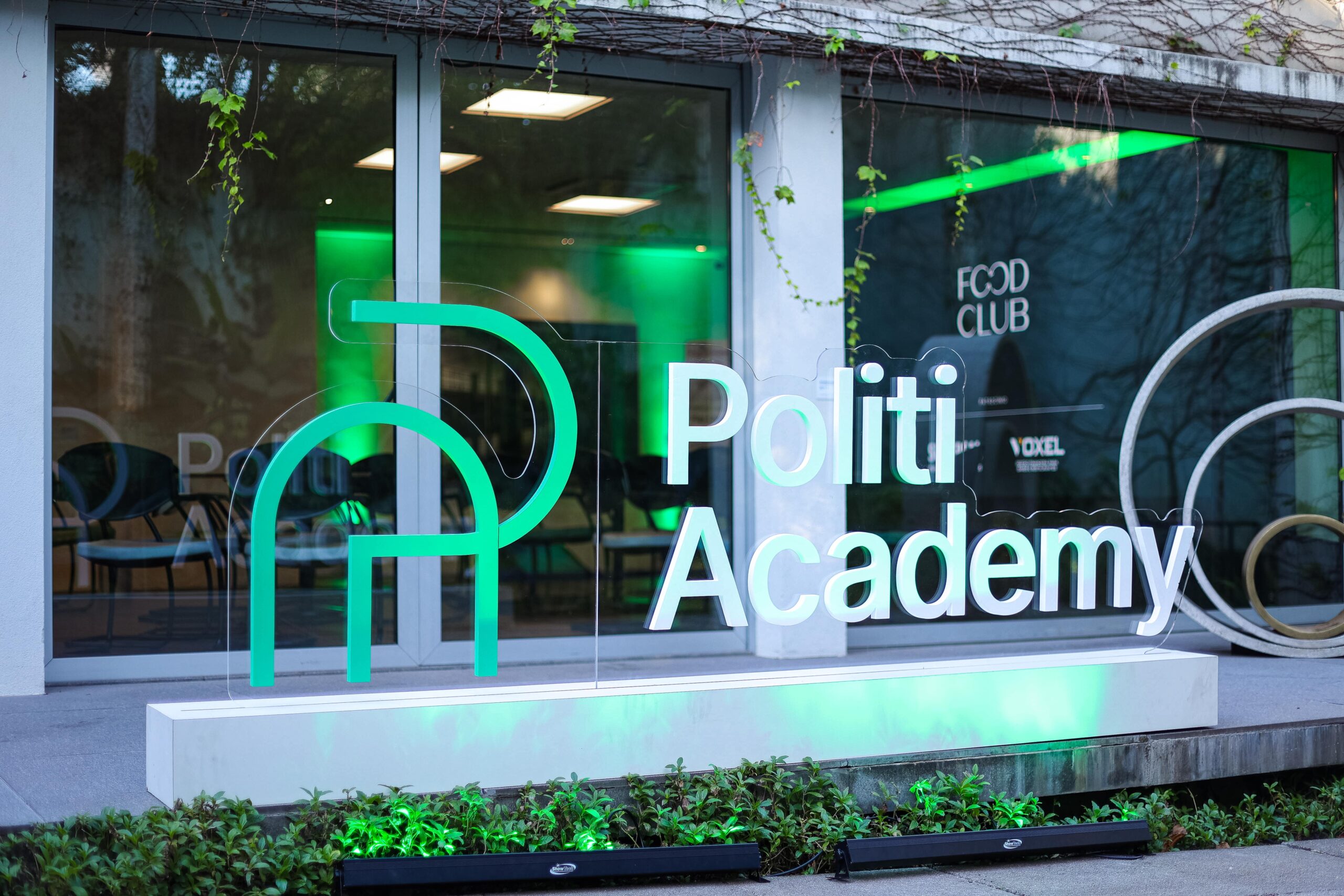 Vista frontal do prédio da Politi Academy com grandes janelas de vidro e uma placa verde e branca com os dizeres "Politi Academy" perto da entrada, ao lado do Food Club.