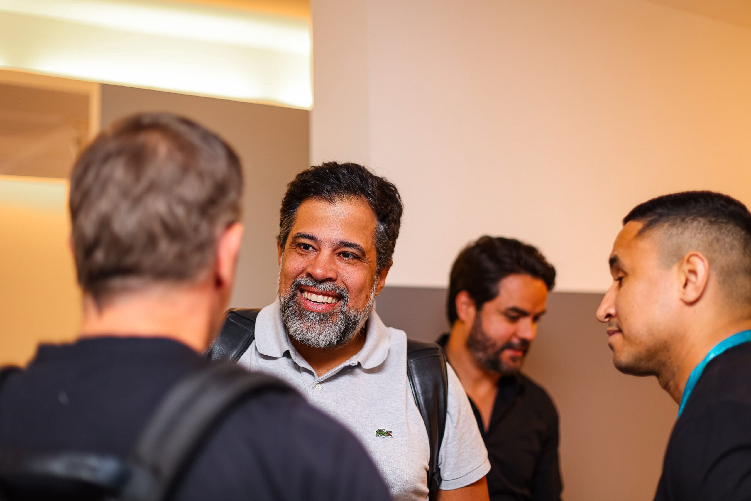 Quatro homens conversam em uma sala bem iluminada. Um deles, com cabelos grisalhos e barba, sorri calorosamente enquanto veste uma camisa polo de cor clara. O ambiente parece um clube exclusivo onde a boa comida pode estar logo ali na esquina.