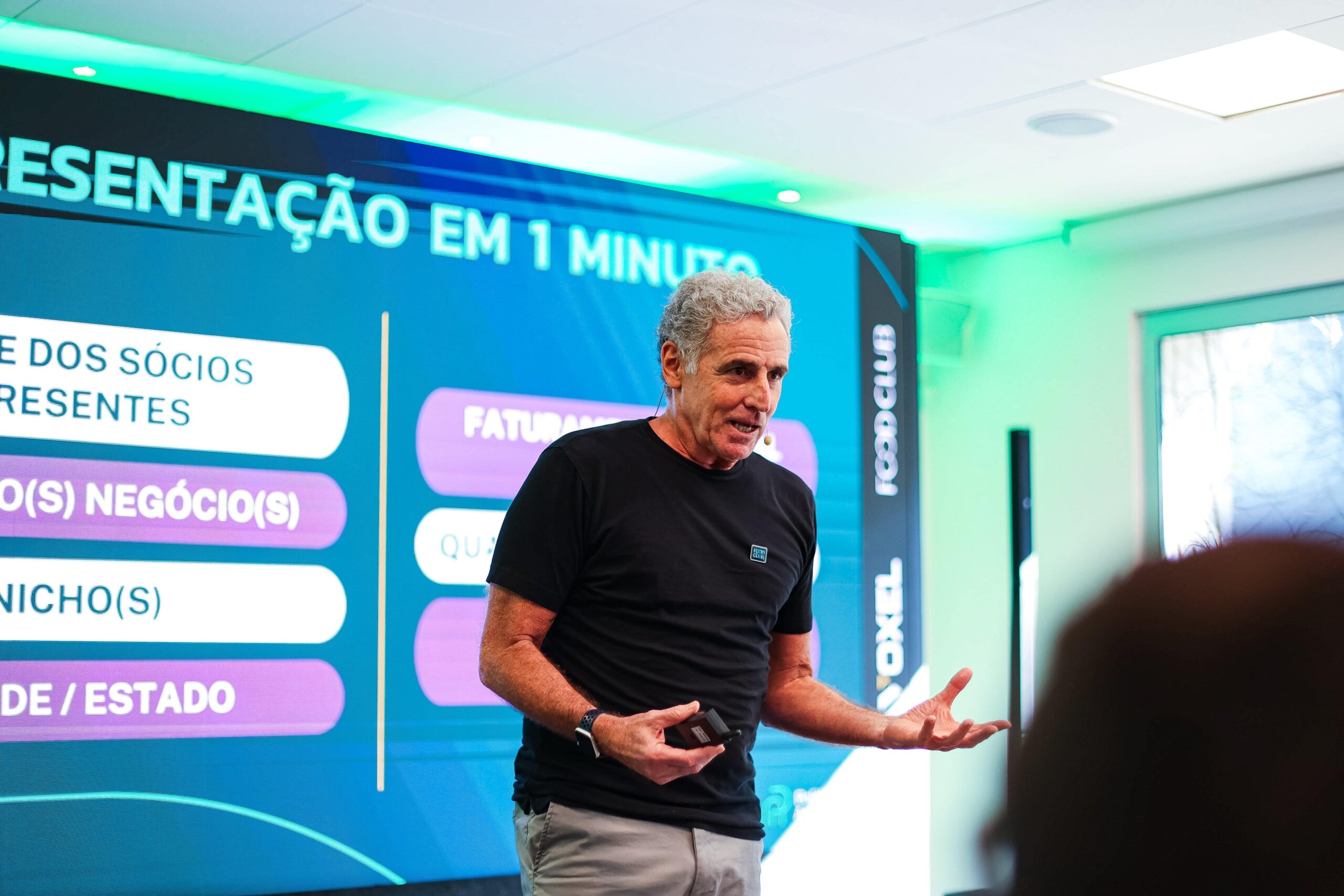 Uma pessoa com cabelos grisalhos está fazendo uma apresentação em uma sala moderna com uma grande tela digital atrás dela exibindo texto em português. O apresentador, gesticulando com as mãos, discute os próximos eventos do clube de alimentação.