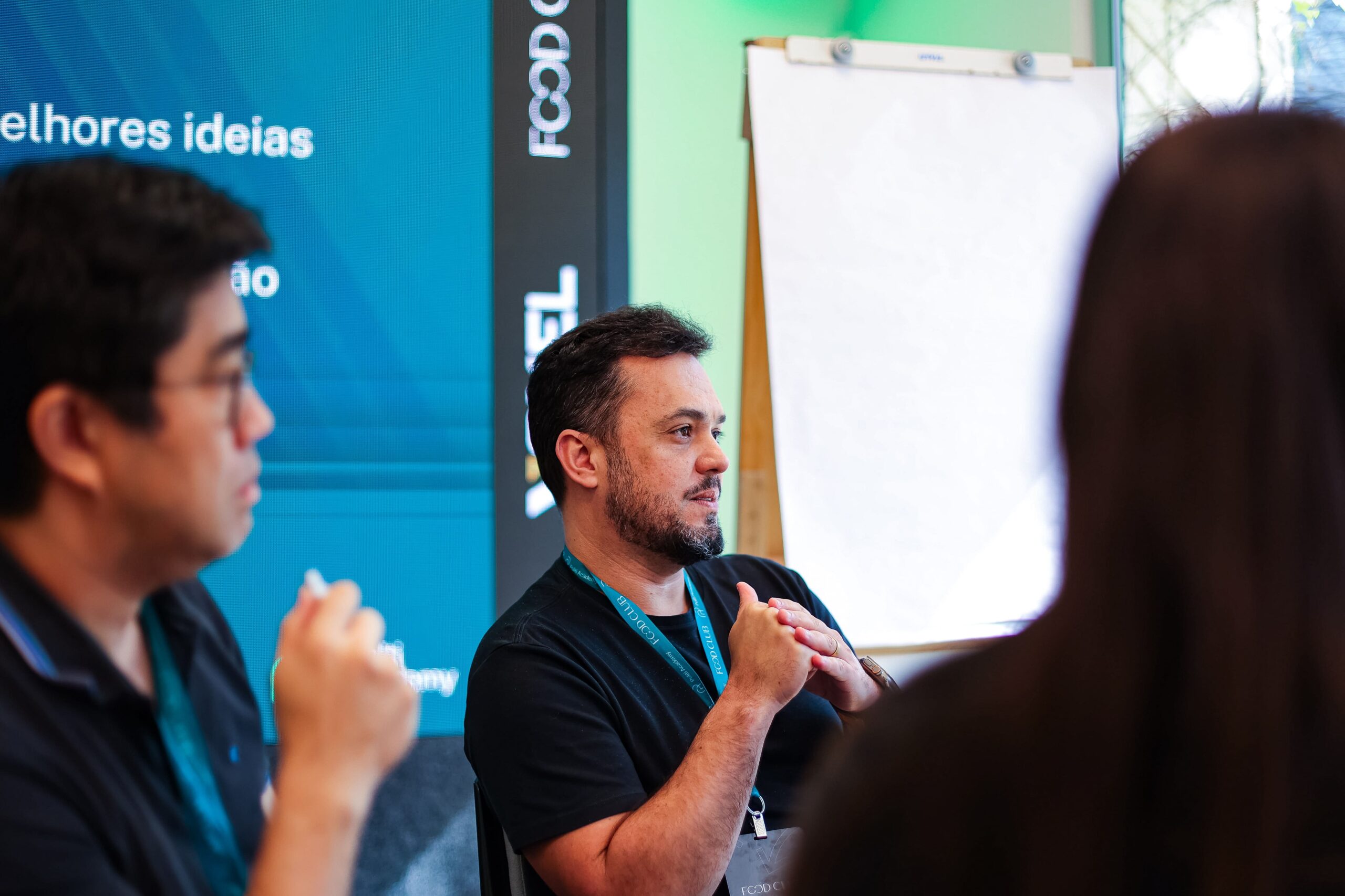 Um homem está sentado em uma mesa de discussão, falando enquanto segura uma caneta. Um flip chart é visível no fundo, e outra pessoa está desfocada no primeiro plano. Uma tela com texto é parcialmente visível. É como se ele estivesse apresentando ideias para o novo menu de comida do clube.
