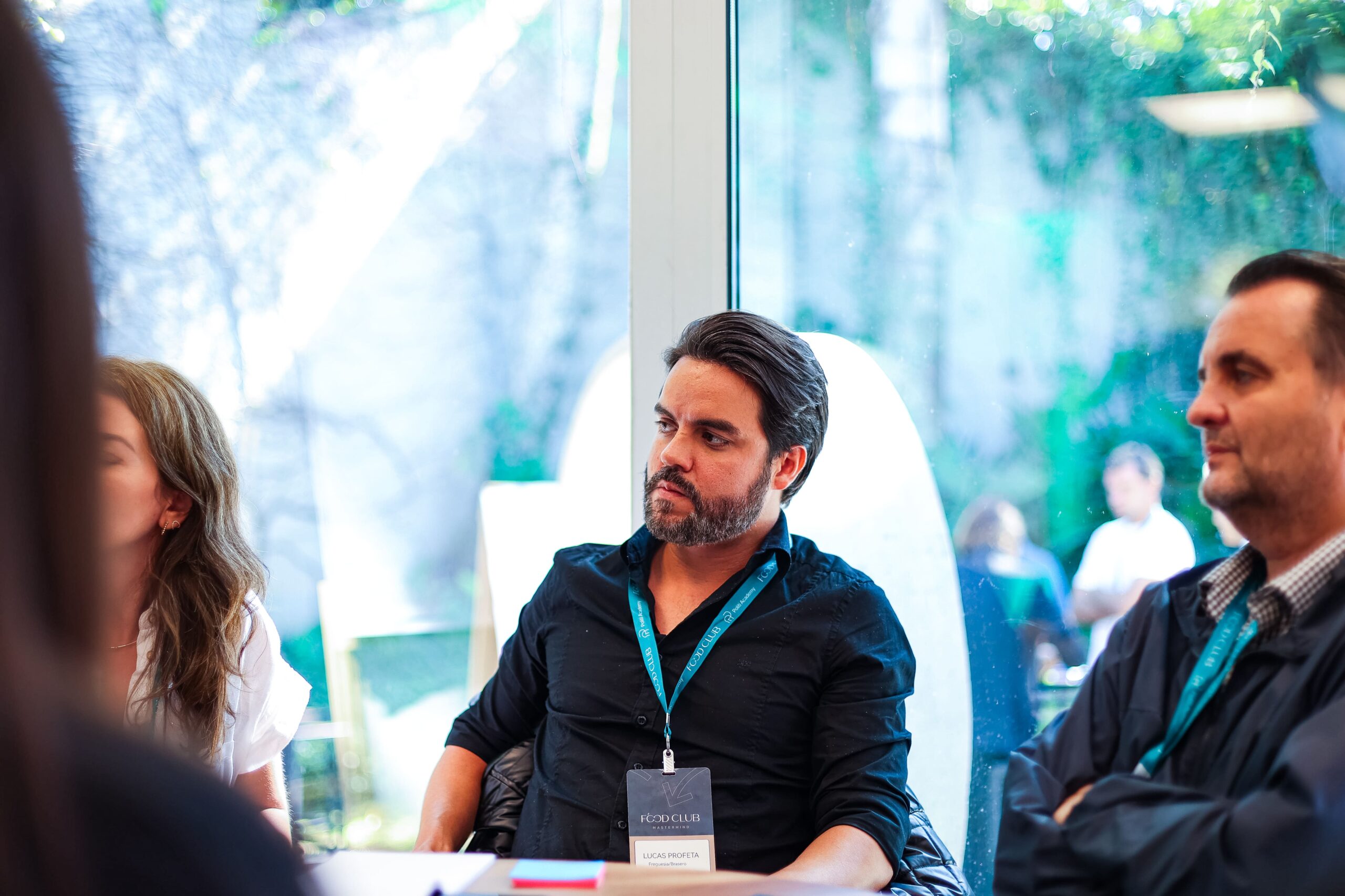 Um grupo de pessoas senta-se ao redor de uma mesa em uma sala bem iluminada com uma grande janela mostrando a vegetação do lado de fora. Um homem de camisa preta e cordão, claramente parte do Food Club, está em foco, olhando atentamente para sua esquerda.