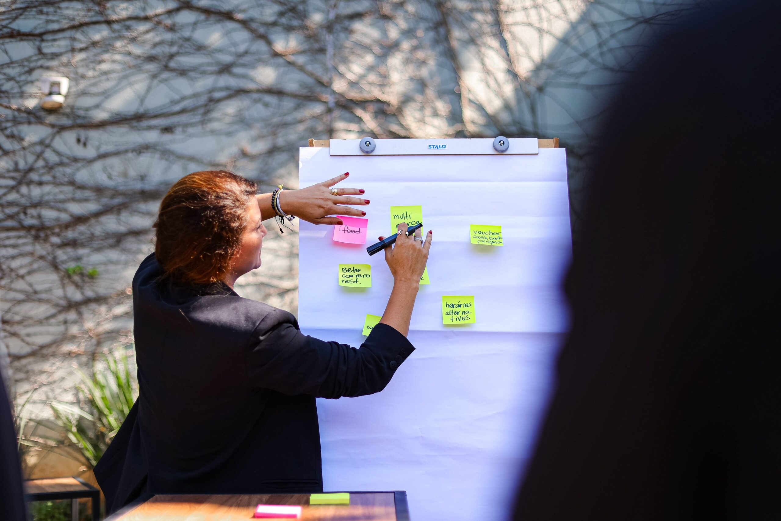Uma pessoa está em frente a um flip chart ao ar livre, colocando notas adesivas e escrevendo com um marcador, organizando ideias para a próxima reunião do Food Club.
