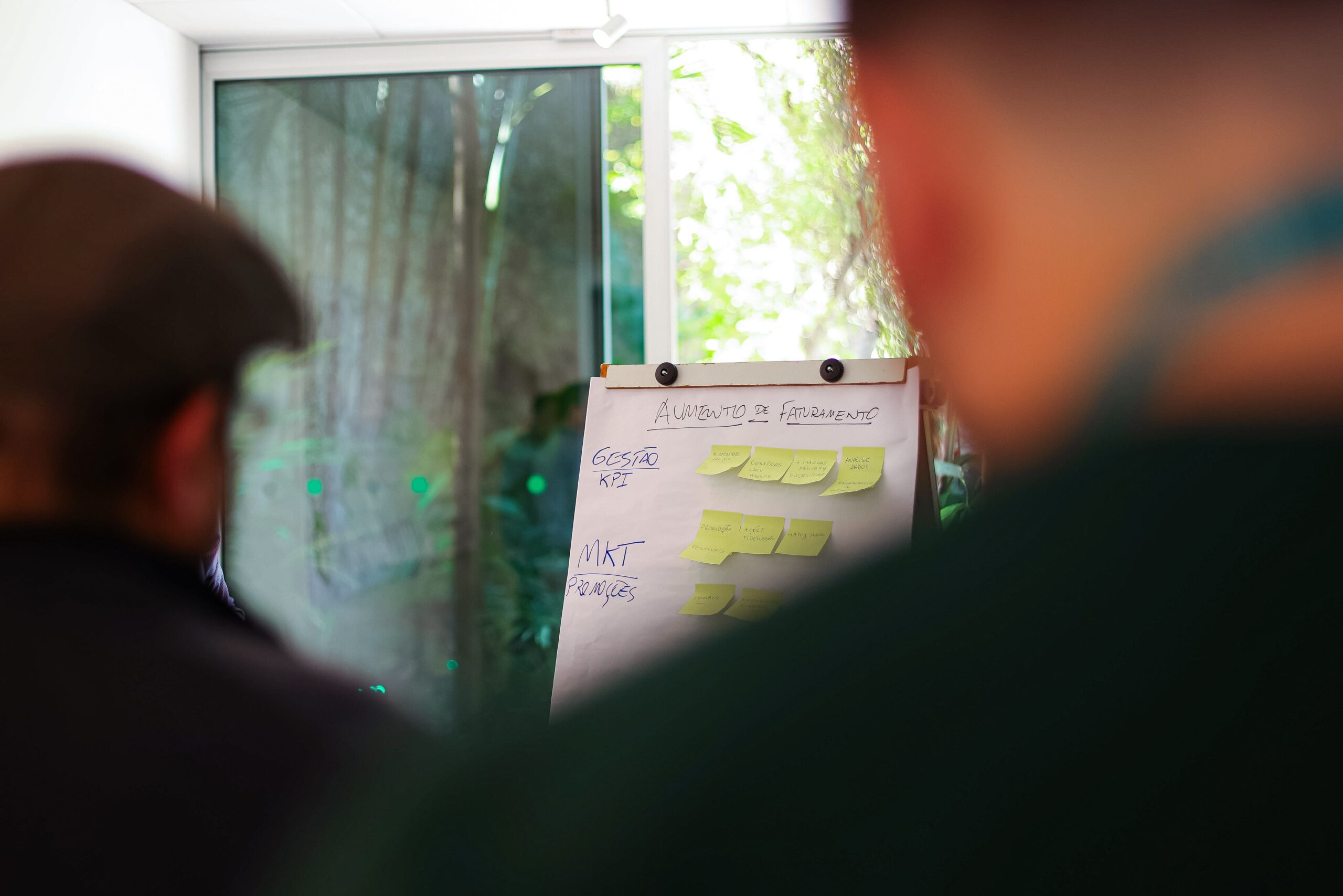 Duas pessoas observam um quadro branco com post-its e texto escrito à mão em um ambiente interno, fazendo brainstorming de ideias para seu Food Club. Uma janela com vegetação do lado de fora está ao fundo, adicionando um toque de natureza à sua sessão criativa.