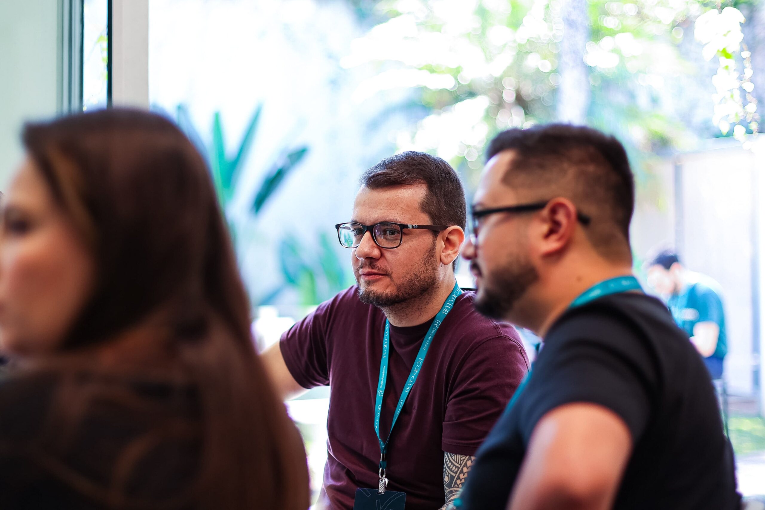 Três indivíduos estão dentro de casa, envolvidos em uma conversa. Dois homens, presumivelmente membros de um clube, estão em foco; um está usando óculos e uma camisa marrom, o outro está com uma camisa preta. Outra pessoa está desfocada no primeiro plano.
