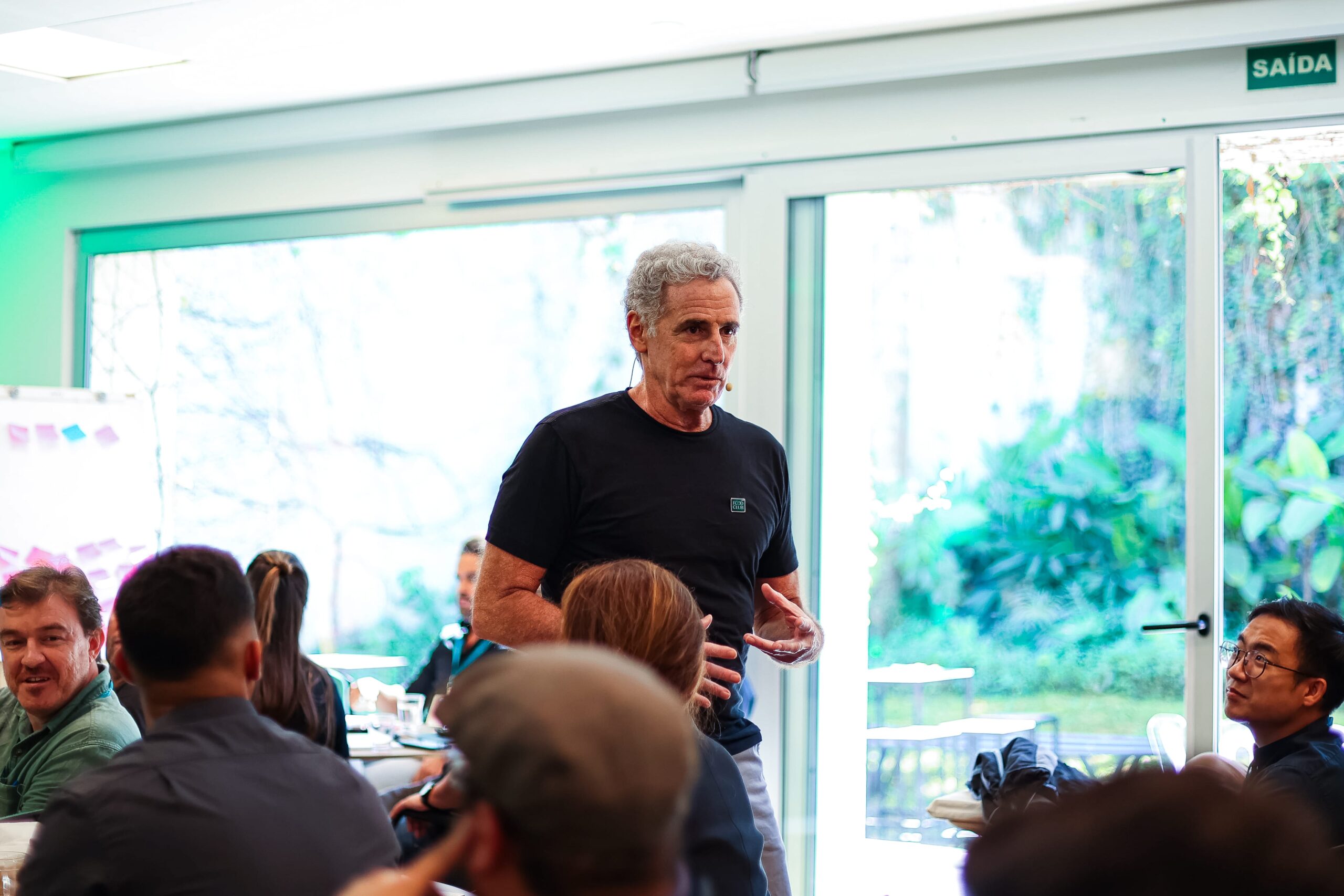 Um homem de camisa preta fala para uma plateia sentada ao redor de mesas em uma sala bem iluminada com grandes janelas, criando a atmosfera de um clube exclusivo.