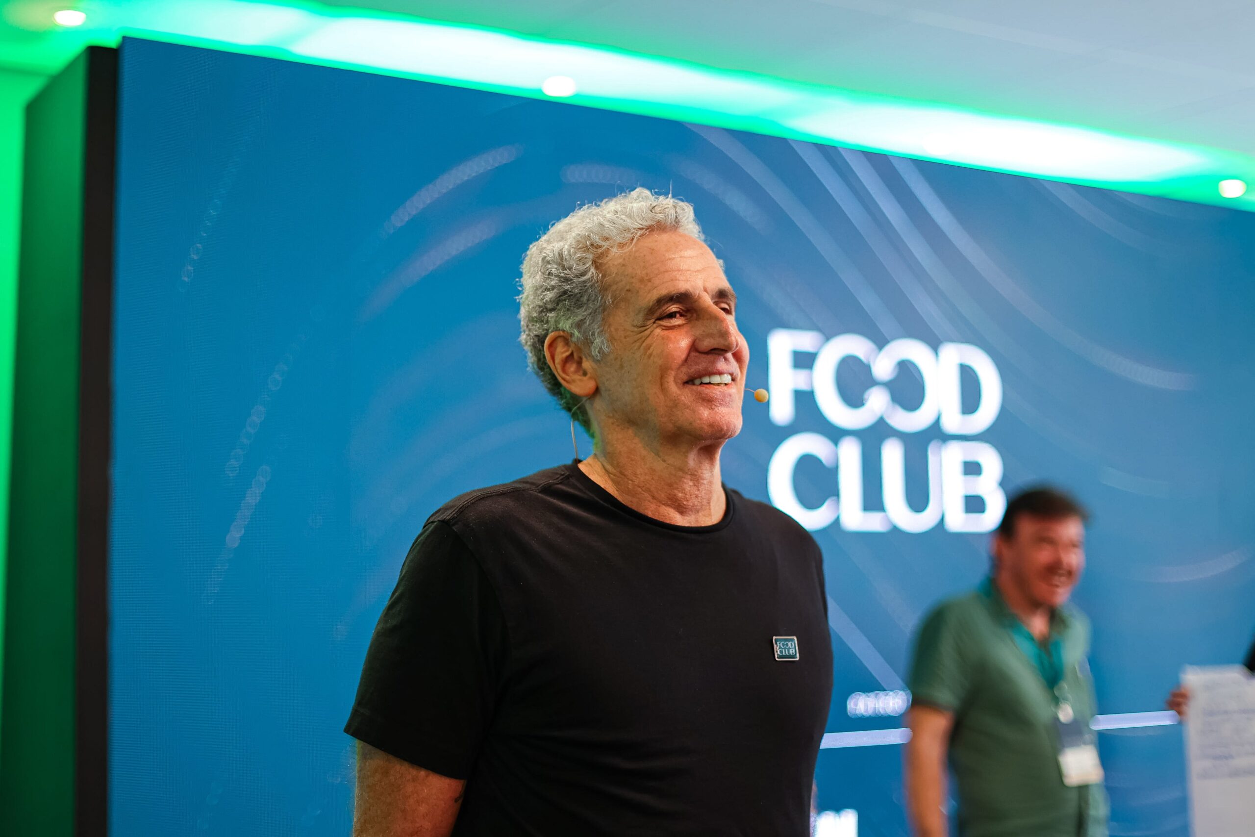 Um homem com cabelos grisalhos e cacheados está em pé em frente a um fundo azul adornado com as palavras "FOOD CLUB". Outro homem ao fundo está sorrindo.