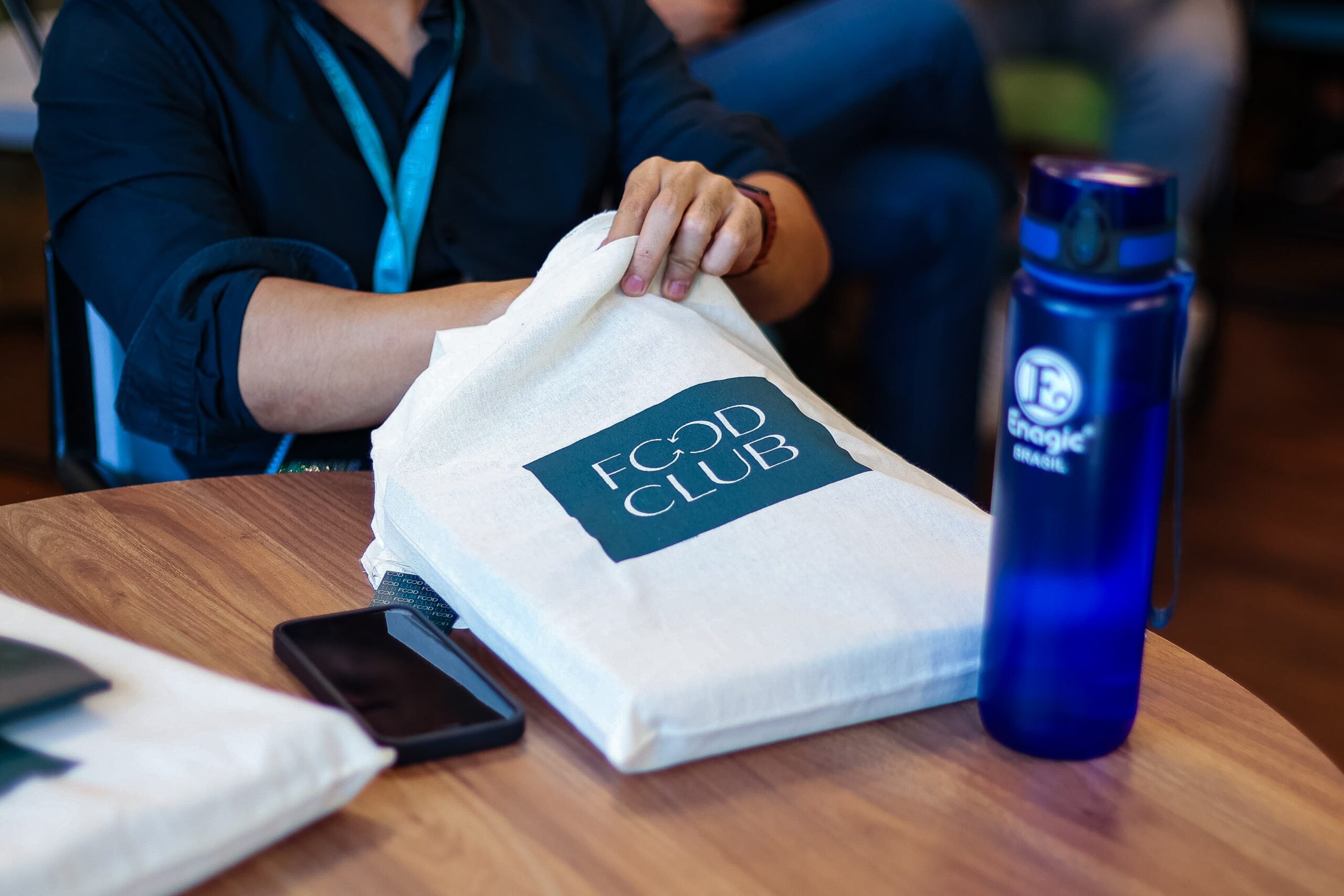 Pessoa de camisa escura pegando uma sacola branca com a etiqueta "FOOD CLUB" em uma mesa de madeira com uma garrafa de água azul e um smartphone.