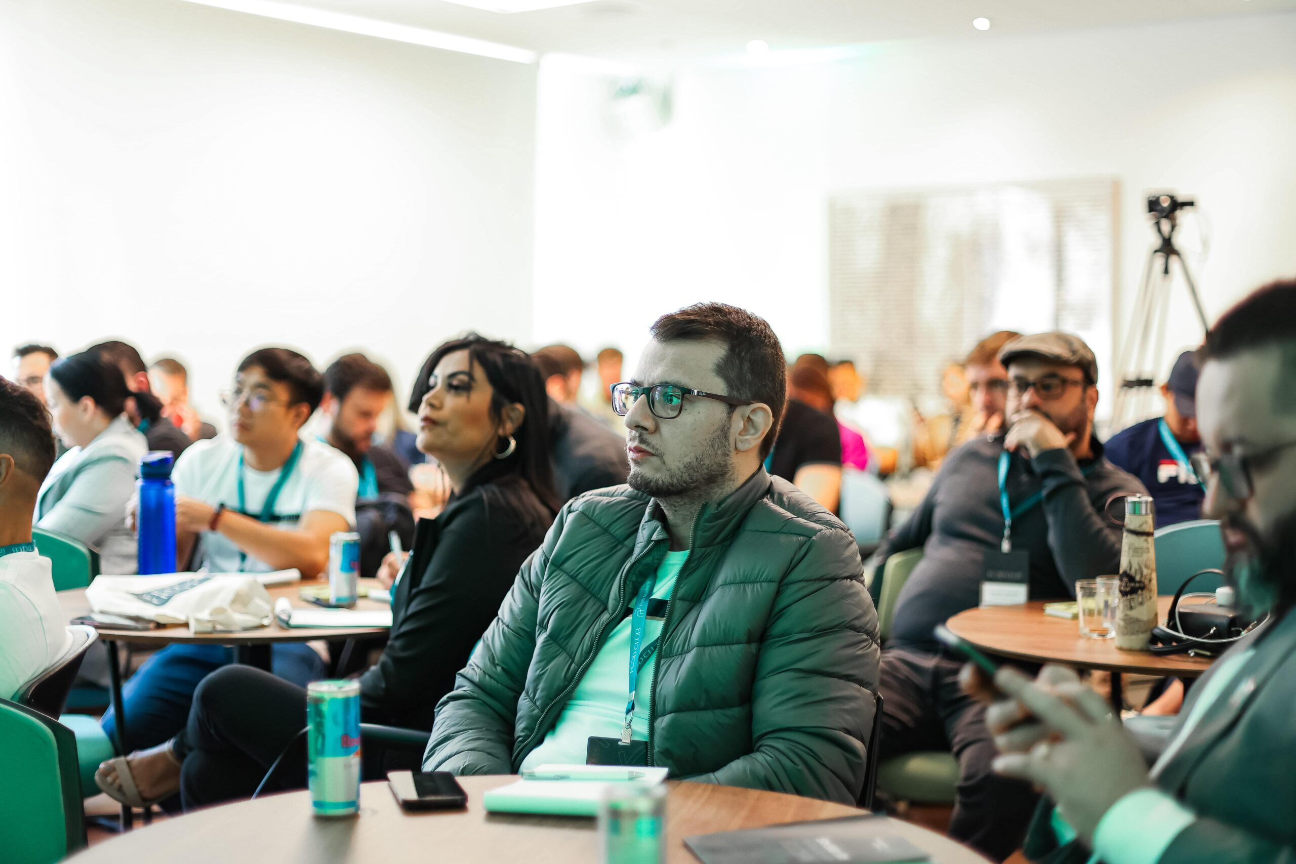 Um grupo de pessoas sentadas em mesas em uma sala de conferências, ouvindo atentamente uma apresentação. Alguns estão tomando notas ou usando seus telefones, enquanto outros conversam sobre as últimas tendências de suas reuniões do Food Club.