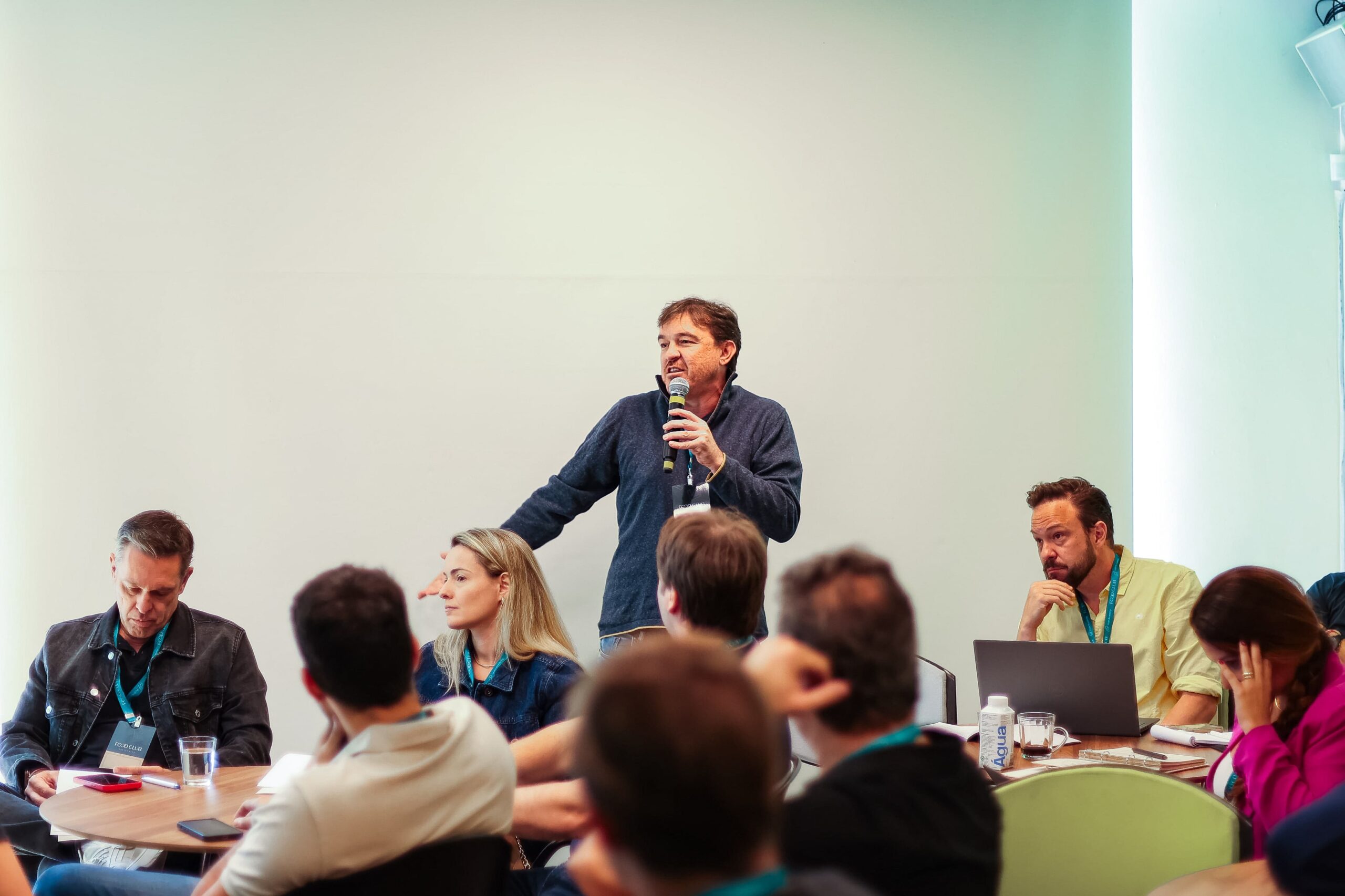 Um homem fala em um microfone enquanto está de pé em uma mesa durante a reunião do Food Club. Várias pessoas sentadas ao redor dele ouvem atentamente, algumas tomando notas e outras olhando para dispositivos eletrônicos.